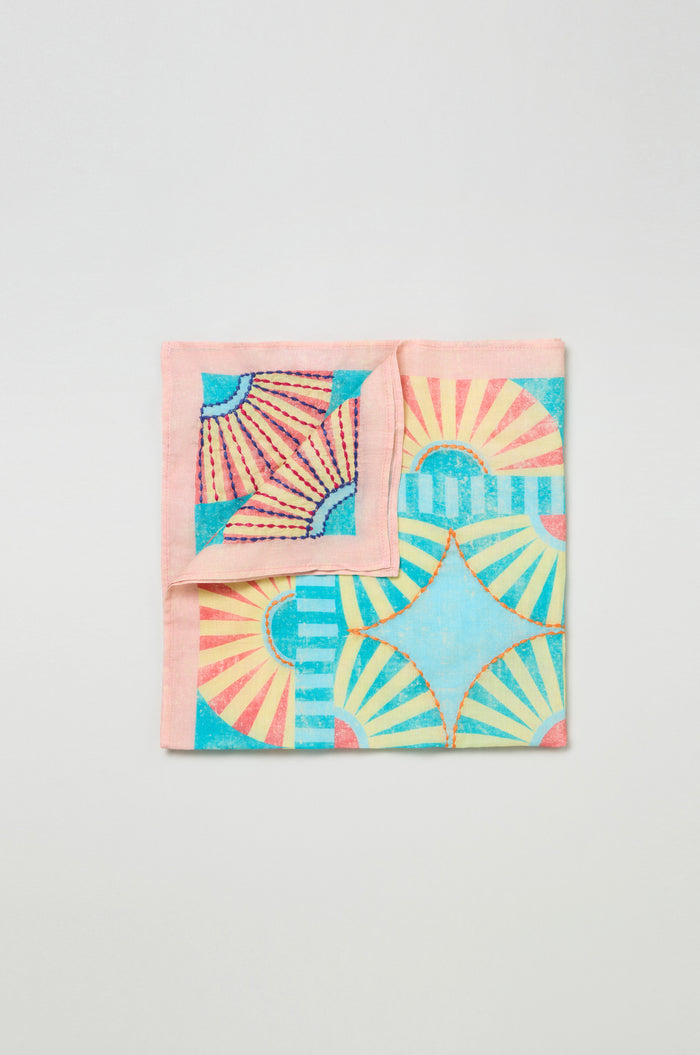The Embroidered Parasols Cotton Bandana is a square cotton accessory with a folded corner, showcasing vibrant geometric sunburst and star designs in pink, blue, yellow, and teal.
