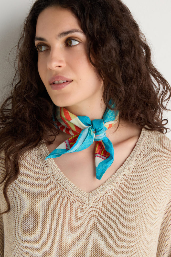 A woman with curly brown hair wears a beige knit top and the Embroidered Parasols Cotton Bandana tied around her neck, gazing slightly to the side against a plain backdrop.