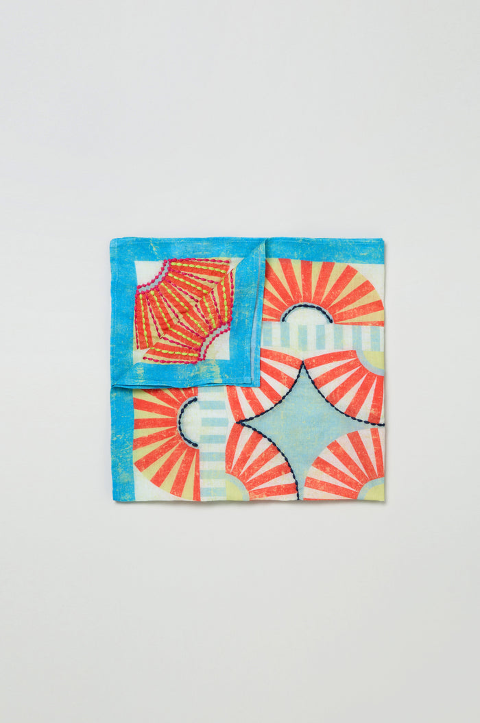 The Embroidered Parasols Cotton Bandana features a colorful geometric design with red sunburst patterns, blue borders, and a central star-shaped cutout, evoking a parasol motif. Displayed on a plain background.
