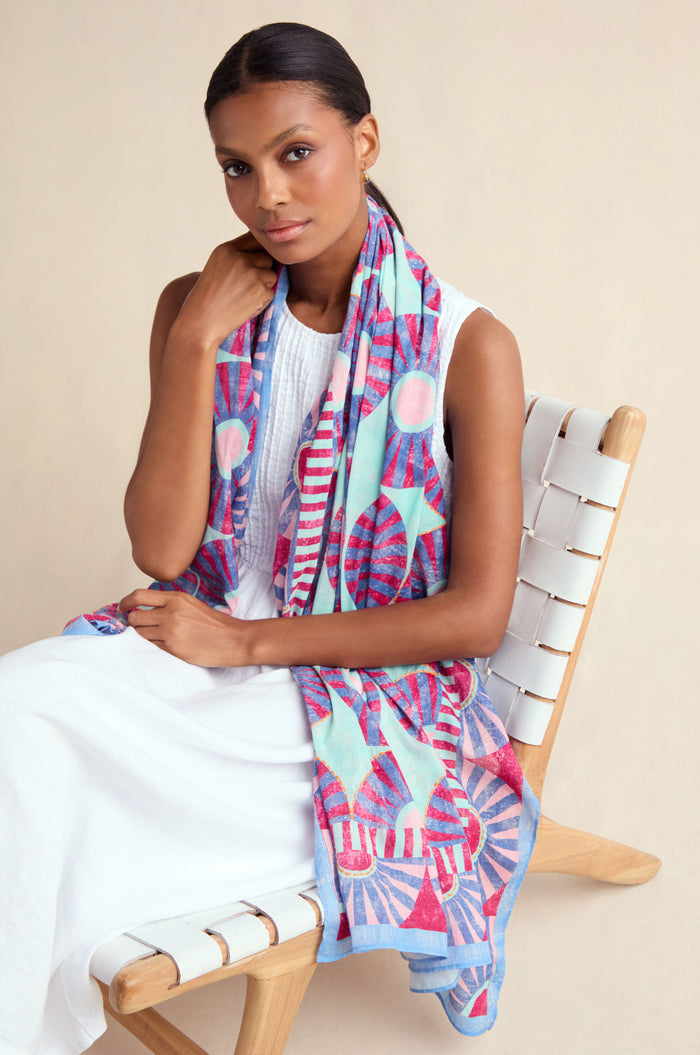 A woman in a white sleeveless dress sits on a light woven chair, draping the Embroidered Parasols Cotton Scarf—featuring a colorful geometric pattern and lightweight cotton—over her shoulders.