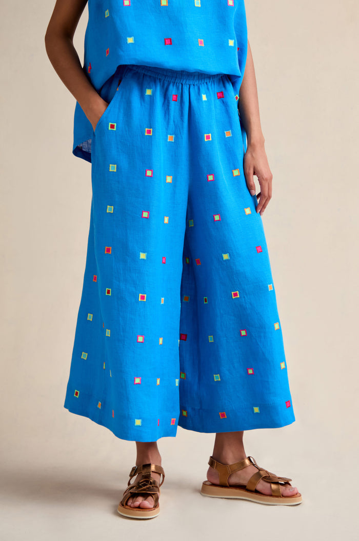 Wearing the Embroidered Squares Linen Wide Trousers in bright blue with a matching cropped top and brown sandals, this person stands against a neutral background.