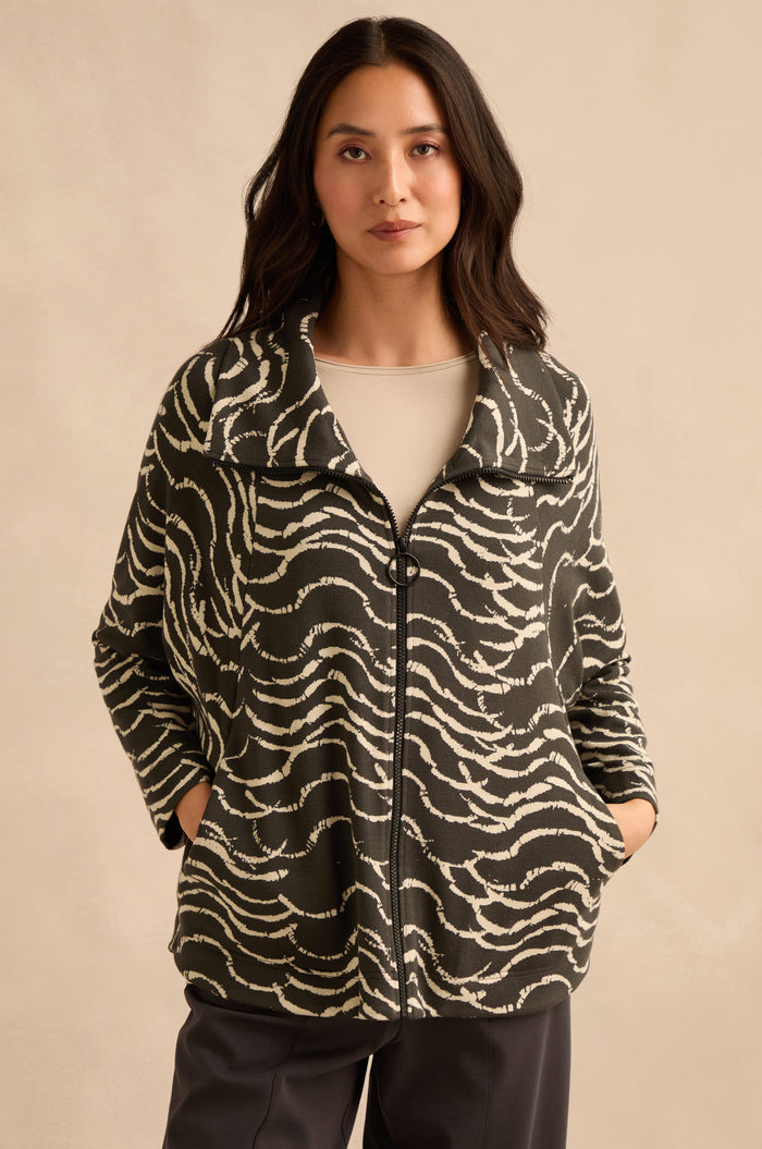 A woman stands with her hands in the pockets of a Dancing Waves Zip Boxy Jacket—featuring a black and white wave motif—paired with a light beige top and dark pants, against a plain beige background.