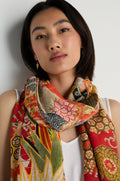 A woman with straight dark hair wears a white top and the Fans Scarf in scarlet and orange, featuring floral and geometric patterns, standing against a plain white background.