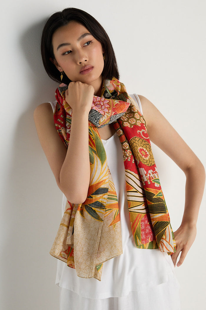 A woman in a white sleeveless top wears the Fans Scarf in scarlet and orange, showcasing floral and geometric patterns on sustainable fabric, standing against a plain light background.