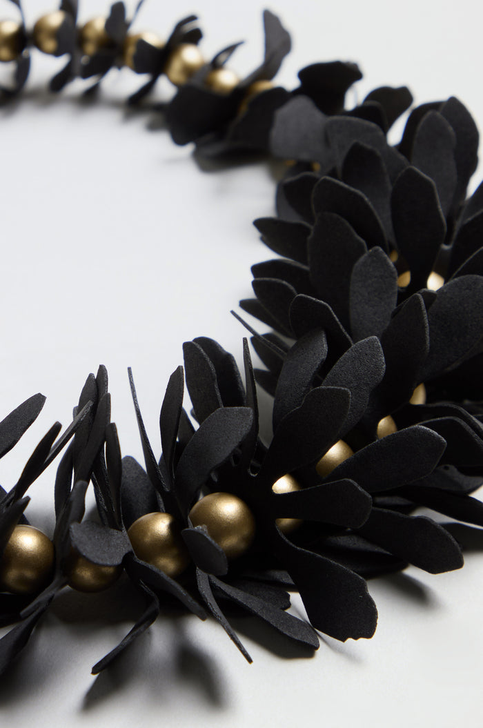 Close-up of the Flower Bead Necklace, featuring black paper leaves and gold accent beads arranged in a circular pattern on a light surface.