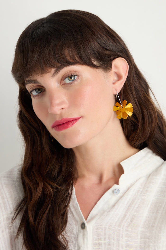 A woman with long brown hair wears a white blouse and Flower Earrings, featuring yellow blooms crafted from anodised aluminium, looking at the camera with a neutral expression.