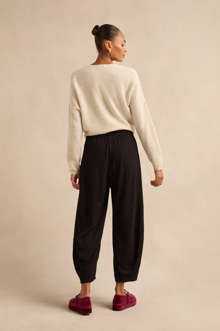 A person stands facing away, wearing a cream sweater, the Fluid Crepe Bubble Trousers in black, and red loafers, against a plain beige background.