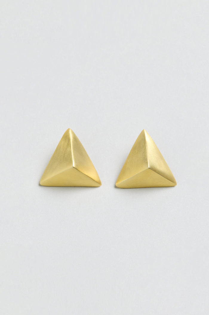 A pair of Folded Triangle Earrings, crafted from recycled brass in a brushed gold finish and featuring a pyramid-shaped geometric design, are displayed against a plain, off-white background.