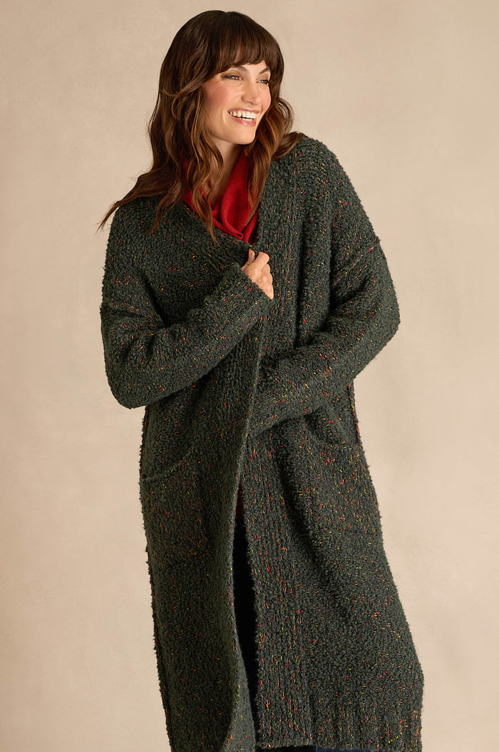A woman stands and smiles against a plain, light-colored background, wearing the Forest Boucle Knit Long Line Speckle Cardi—a long, textured dark green cardigan—layered over a red top.