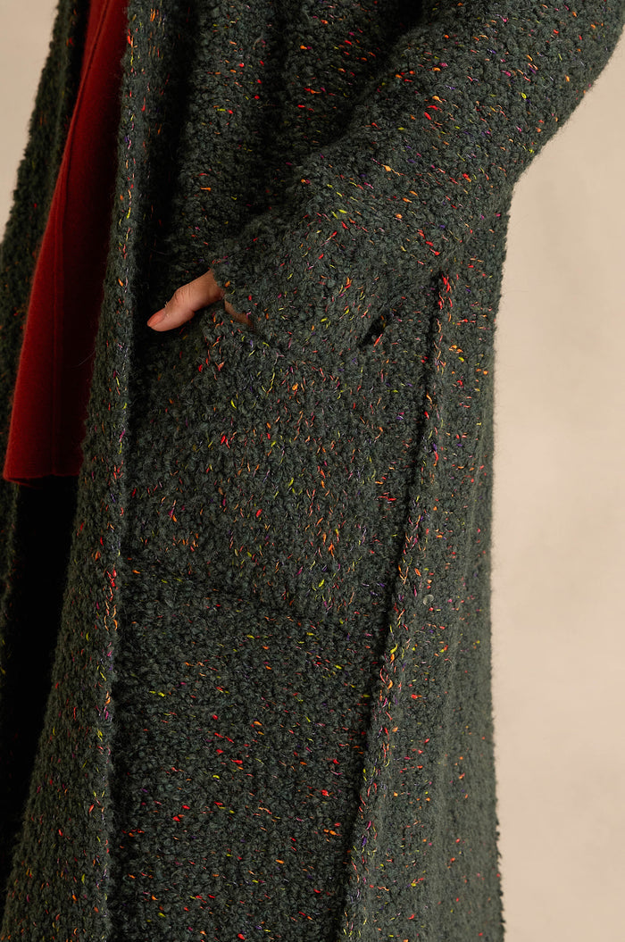 Someone wearing the Forest Boucle Knit Long Line Speckle Cardi, featuring a textured dark green knit with colorful flecks, has their hand tucked into one of its large front pockets.