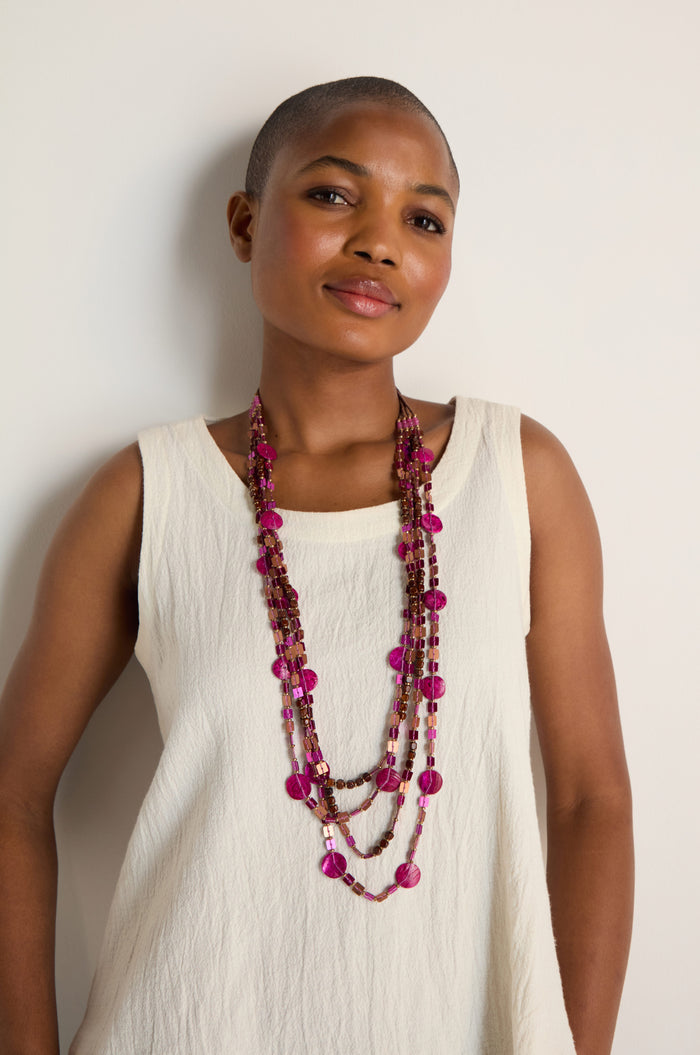 A person with a shaved head wears a sleeveless white top and the Four Strand Sequin Necklace in pink, standing against a plain light background.