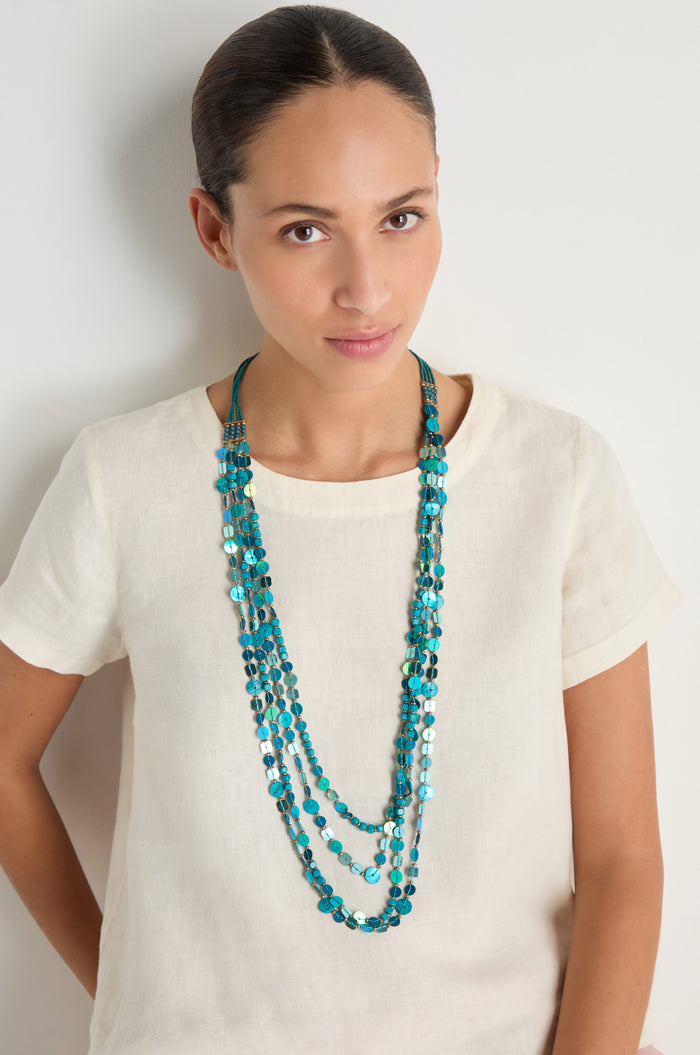 A woman in a cream short-sleeve top and the Four Strand Sequin Necklace with blue beads stands against a plain white background.