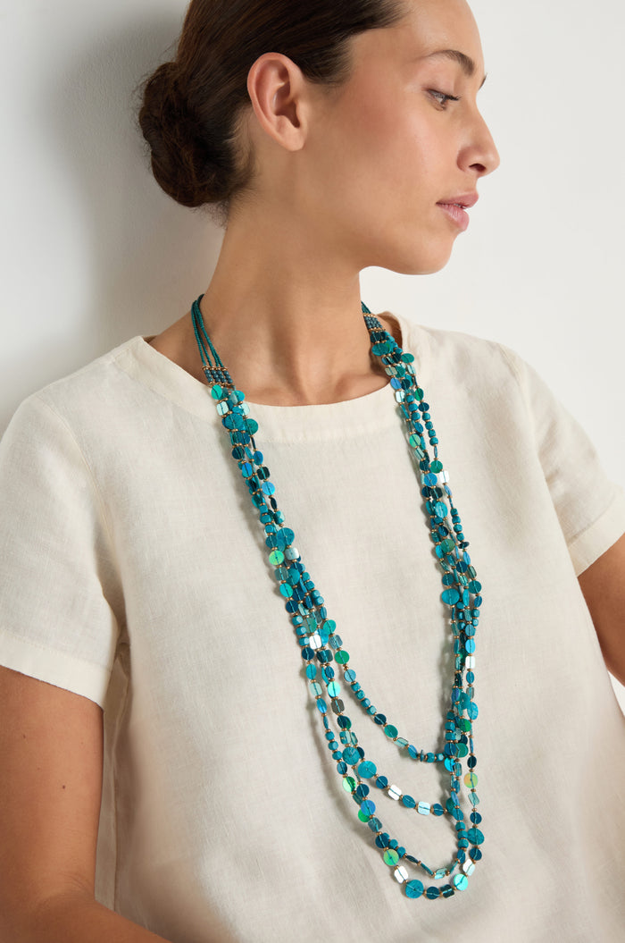 A woman in a cream short-sleeve top wears the Four Strand Sequin Necklace with turquoise beads, looking to the side against a plain white background.