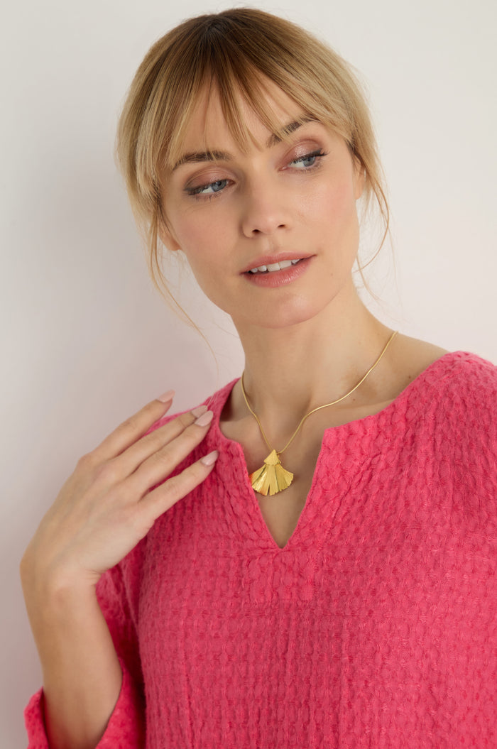 A woman in a textured pink top wears the Gold Vermeil Ginkgo Leaf Pendant necklace, gazing slightly to the side with one hand raised near her neck.