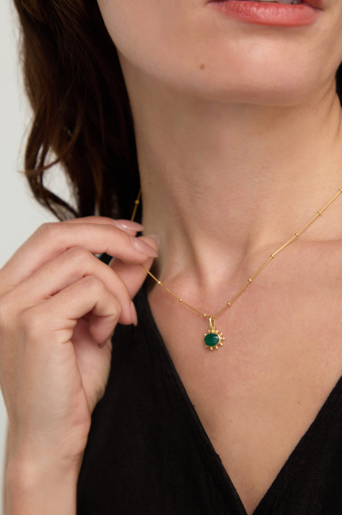 A woman wearing a black top gently holds the Gold Vermeil Green Onyx Necklace, featuring a faceted green onyx pendant, between her fingers.