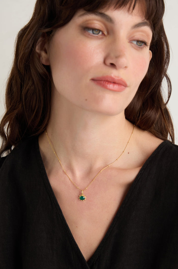 A woman with long brown hair wears a black top and the Gold Vermeil Green Onyx Necklace, featuring a faceted green onyx pendant, as she looks slightly to the side.