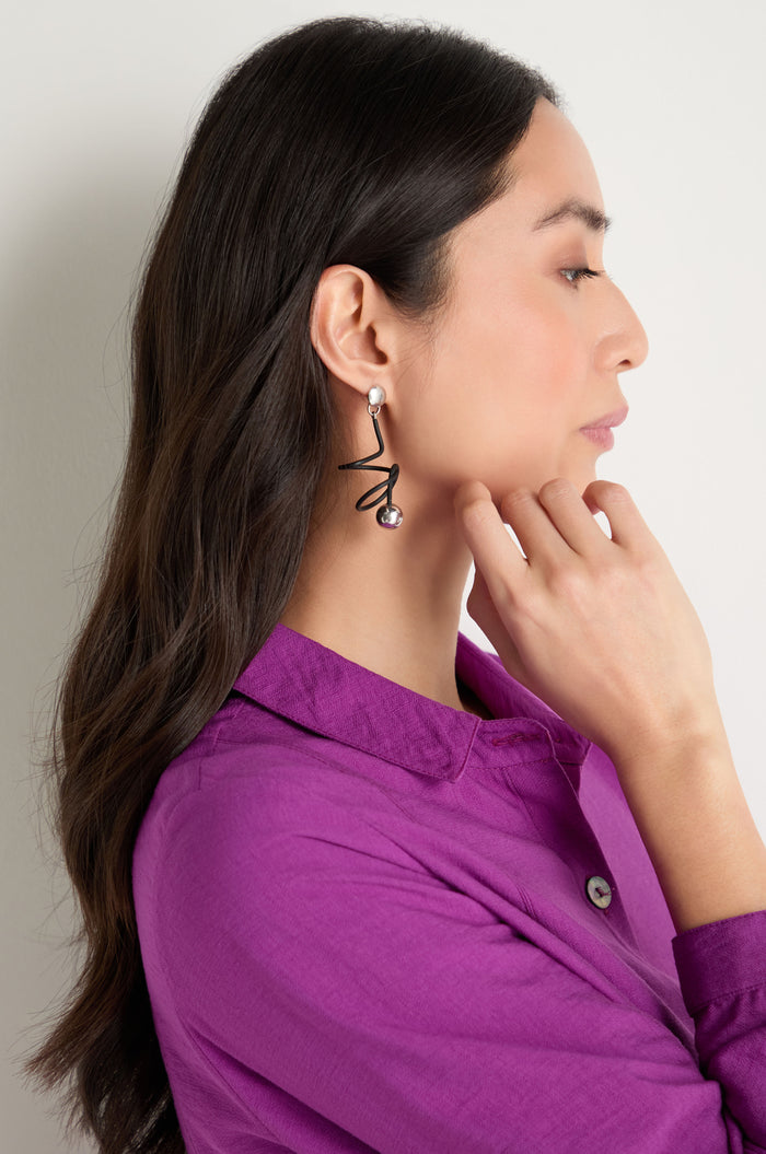 A woman in a purple shirt with long dark hair, shown in profile, thoughtfully touches her chin while wearing contemporary Graphic Squiggle Earrings featuring a bold zigzag design and pearl accents.