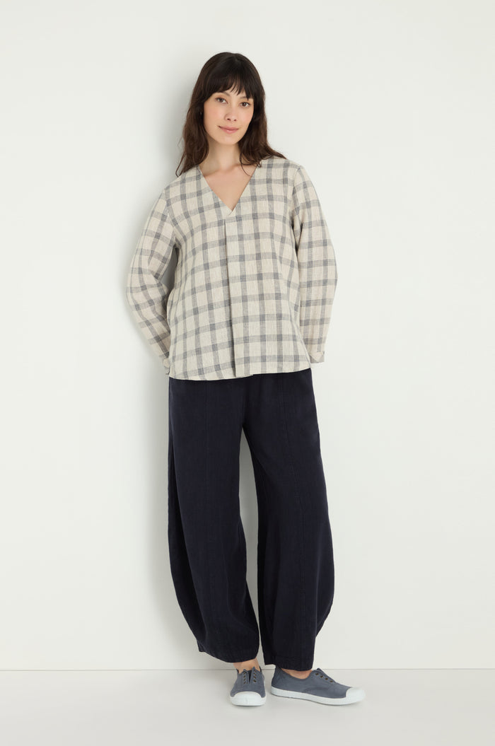 A woman stands against a white wall wearing the Grid Check Linen Top, loose dark pants, and gray sneakers.