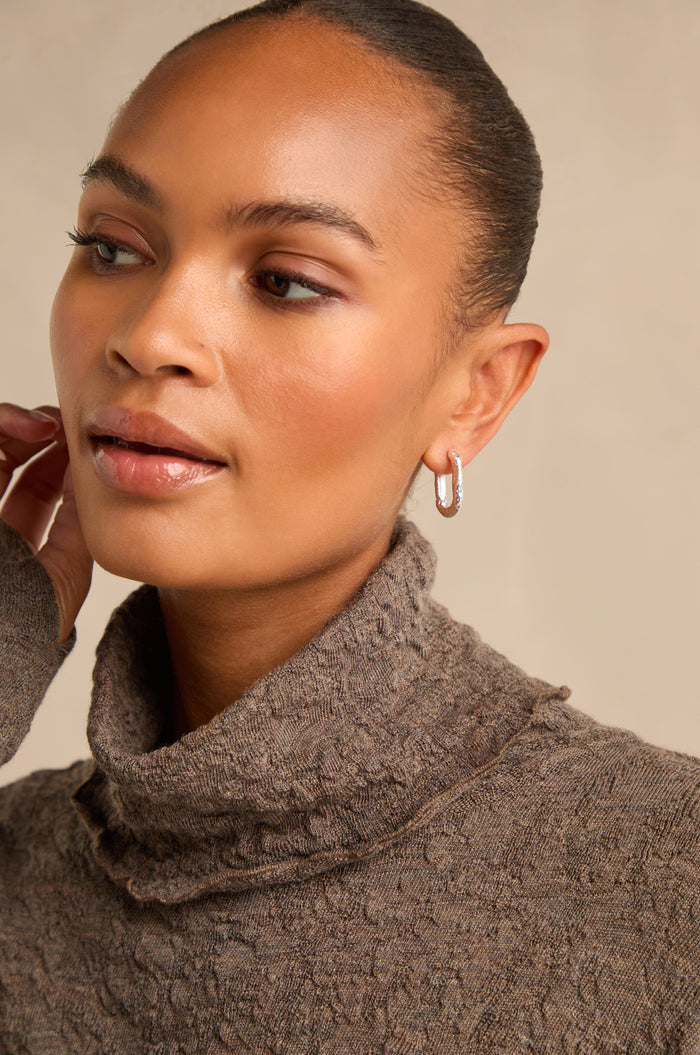 A woman with a neat bun wears the Hammered Hoop Earrings and a textured brown turtleneck sweater, gazing slightly to the side against a neutral background.