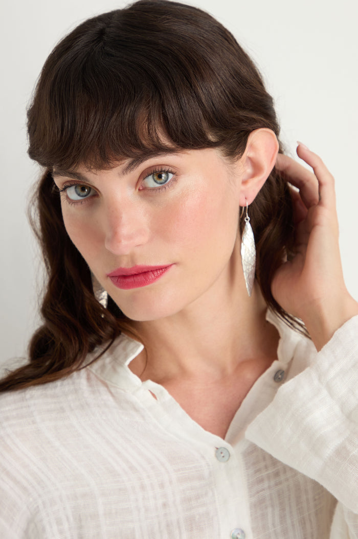 A woman with long brown hair and bangs wears a white button-up shirt and Hammered Silver Leaf Earrings, looking at the camera while touching her hair.