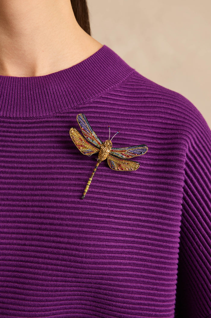 A person wearing a ribbed purple sweater with the Hand Beaded Hawker Dragonfly Brooch pinned to the left side of their chest.