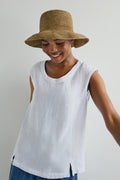 Wearing a Handwoven Raffia Hat, a sleeveless white top, and blue pants, a person smiles while glancing down against a plain background.