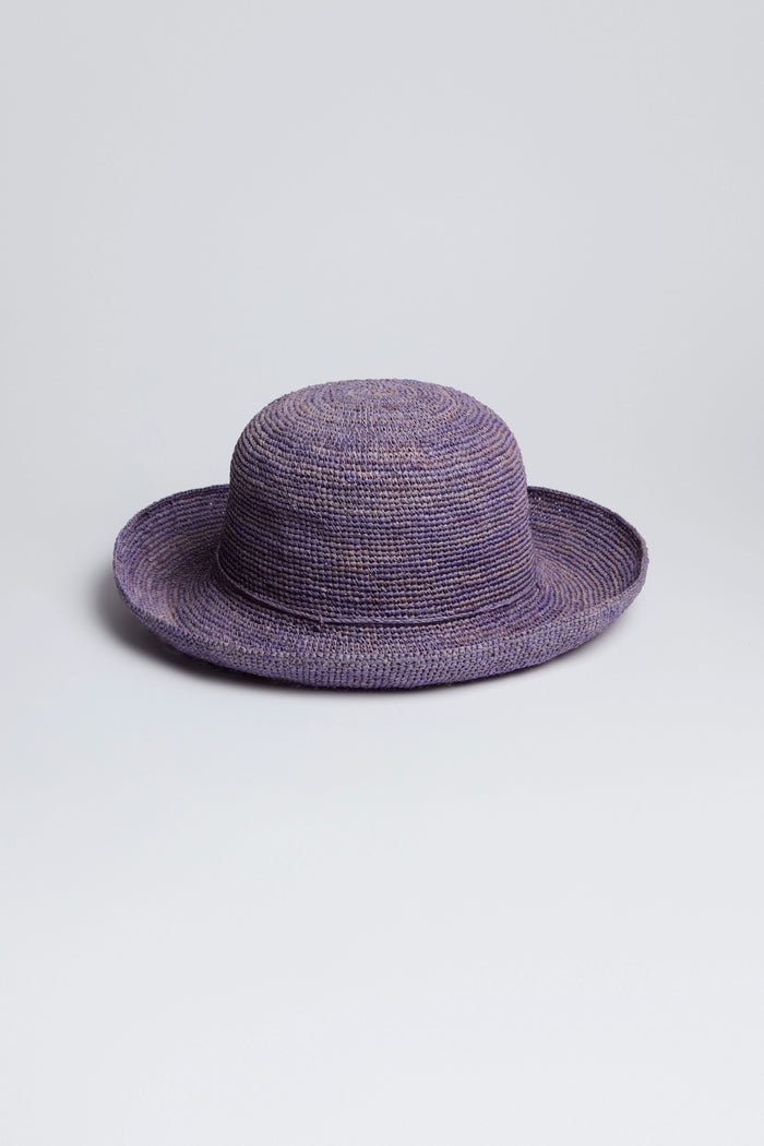 The Handwoven Raffia Hat, crafted in Madagascar, boasts a wide brim and a lovely purple hue, sitting elegantly on any surface.