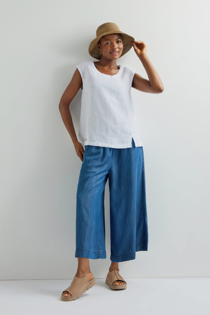 In front of a plain backdrop, someone models a Handwoven Raffia Hat, made with classic Madagascar weaving skills. The ensemble is paired with a white sleeveless top and blue wide-leg pants.