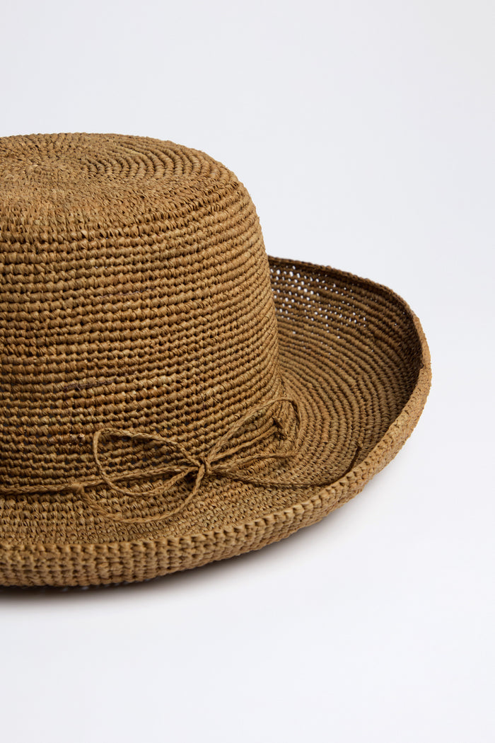 The Handwoven Raffia Hat is a brown straw hat with a round crown and wide brim, crafted using traditional weaving techniques in Madagascar. A small bow detail accents one side.