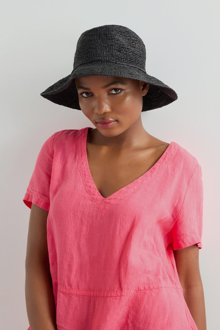 A person in a pink top against a plain background wears a Handwoven Raffia Hat, wide-brimmed and black, crafted with traditional Madagascar weaving techniques, adding an exotic flair to their look.