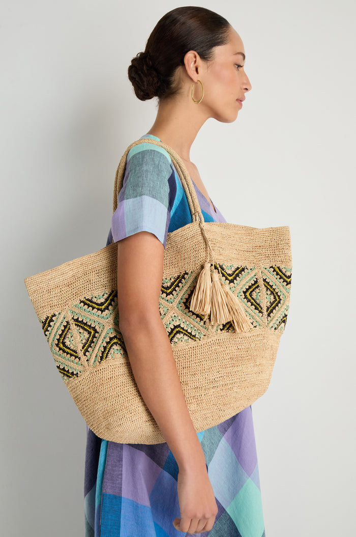 A woman in a multicolored checkered dress carries the Handwoven Raffia Mosaic Bag, featuring bold geometric patterns and tassel details, as she stands against a plain light background.