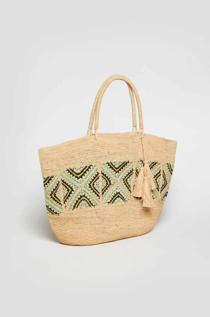 The Handwoven Raffia Mosaic Bag features two handles, a green and black geometric pattern, and two tassels attached to one handle.