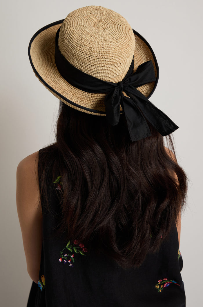 A person with long dark hair, seen from behind, wears the Handwoven Raffia Ribbon Hat—a wide-brim hat with a black ribbon from Madagascar—paired with a black sleeveless top featuring embroidered flowers.