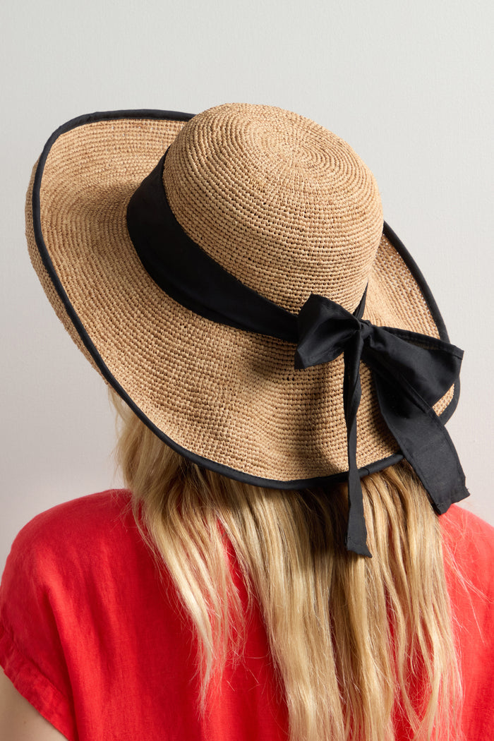 A person with long blonde hair, wearing a Handwoven Ribbon Raffia Hat with a black ribbon, is seen from behind in a red top. The wide-brimmed hat offers excellent sun protection with its artisanal design.