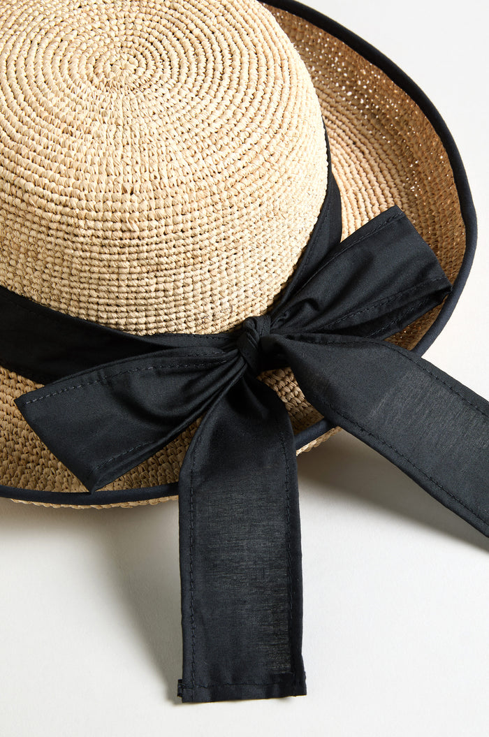 The Handwoven Raffia Ribbon Hat features a wide brim and a large black fabric ribbon bow tied around the crown.