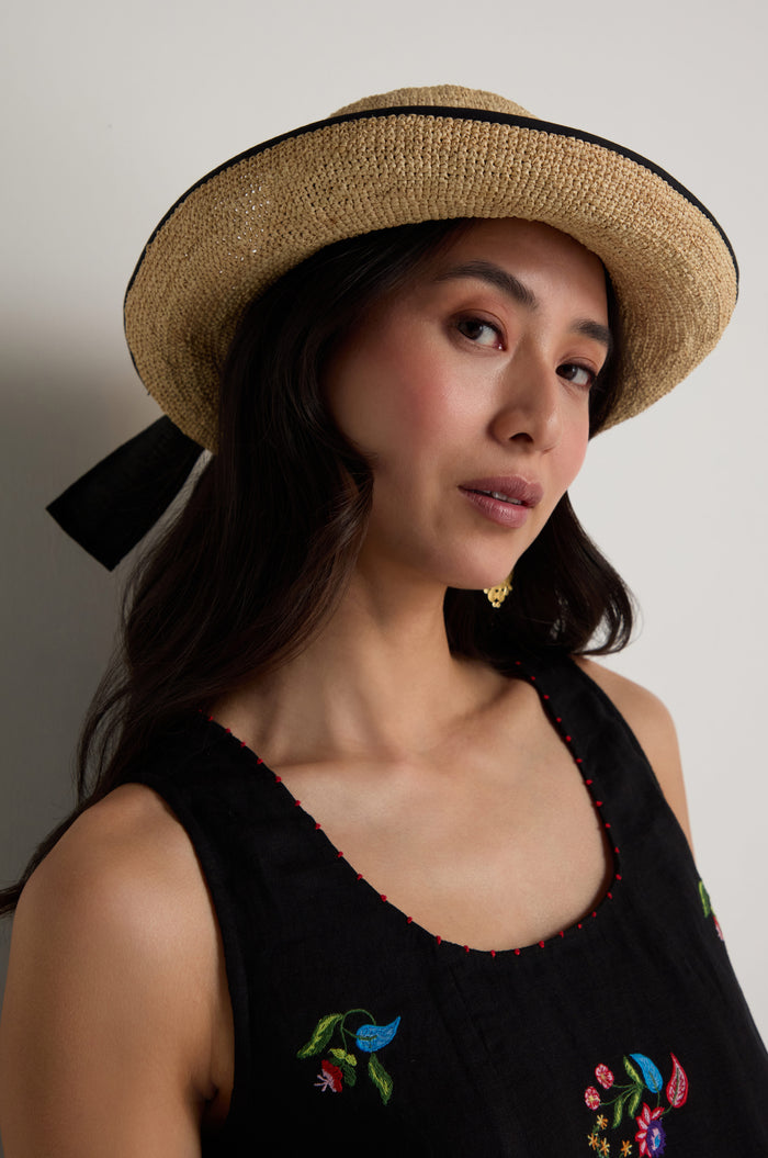 A woman models the Handwoven Raffia Ribbon Hat, crafted in Madagascar with a wide brim and black ribbon, paired with a sleeveless black dress with colorful embroidery, against a plain light background for an elegant look.