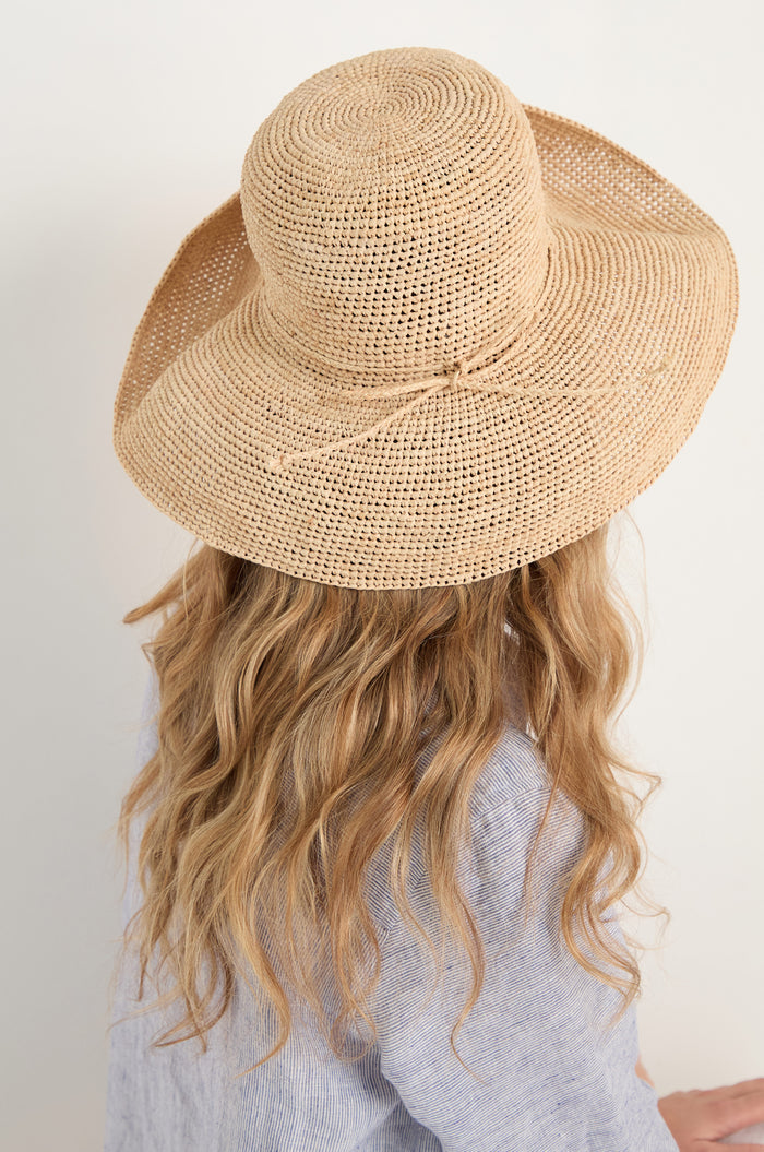 A person with long wavy blonde hair, seen from behind, wears a Handwoven Raffia Tie Hat and a light blue shirt.