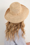 A person with long wavy blonde hair, seen from behind, wears a Handwoven Raffia Tie Hat and a light blue shirt.