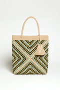 The Handwoven Raffia Tote Bag features a zigzag pattern in tan, black, and light blue, two handles, and a large tassel. Crafted in Madagascar, this artisan bag brings stylish flair to your look.