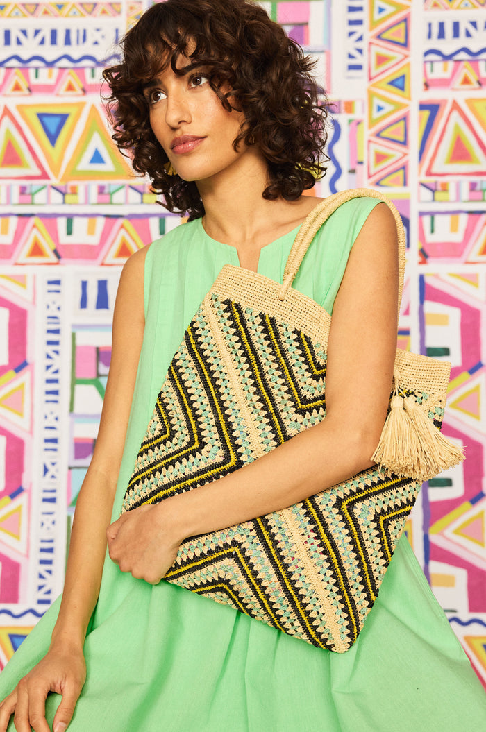 A woman in a sleeveless light green dress holds the Handwoven Raffia Tote Bag with zigzag patterns and tassels, standing before a vibrant geometric-patterned background.
