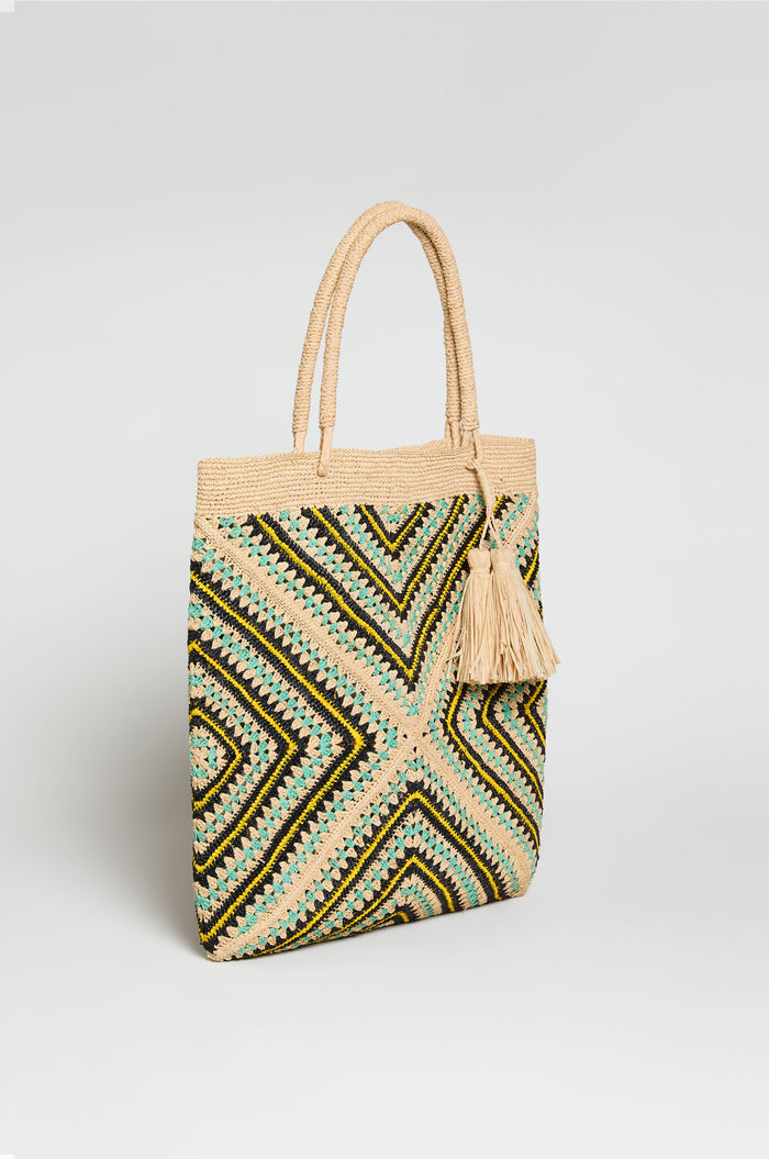 The Handwoven Raffia Tote Bag features artisan craftsmanship, geometric black, yellow, and green patterns, two handles, and a large beige tassel.