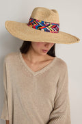 Wearing a beige knit top and the Handwoven Raffia Western Hat featuring a colorful geometric band, this person looks down against a plain background, blending effortless style with sun protection.