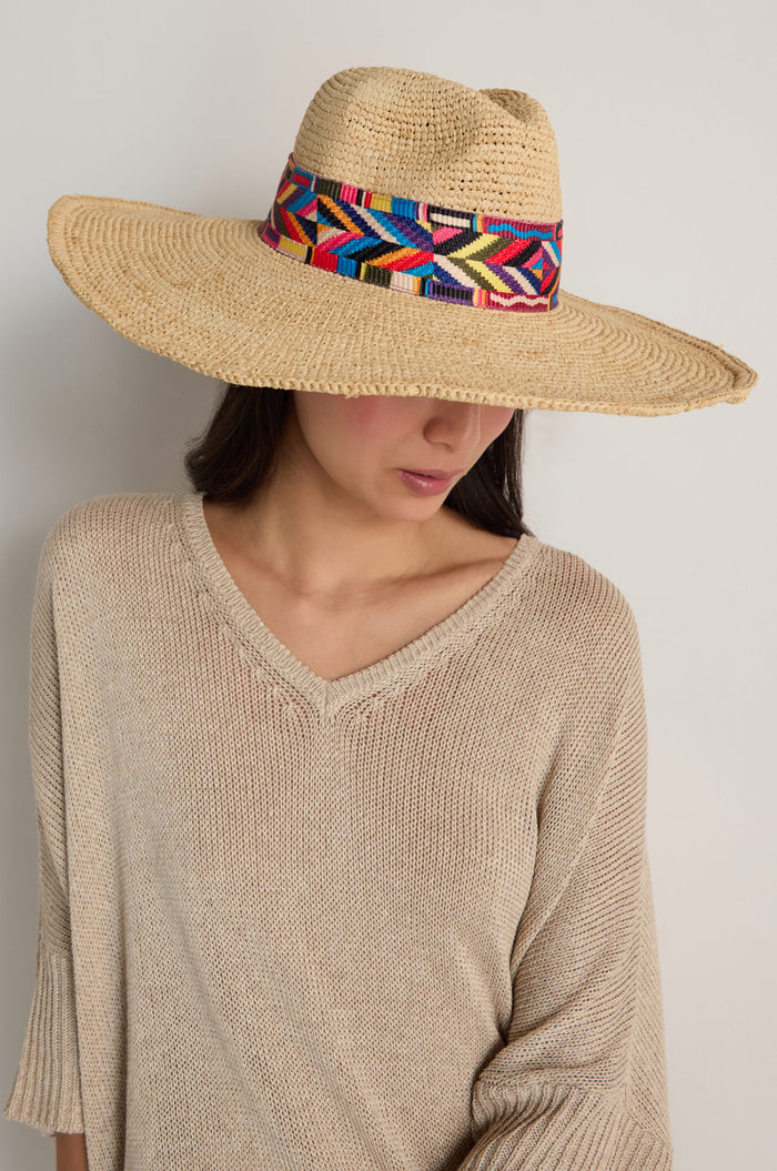 Wearing a beige knit top and the Handwoven Raffia Western Hat featuring a colorful geometric band, this person looks down against a plain background, blending effortless style with sun protection.
