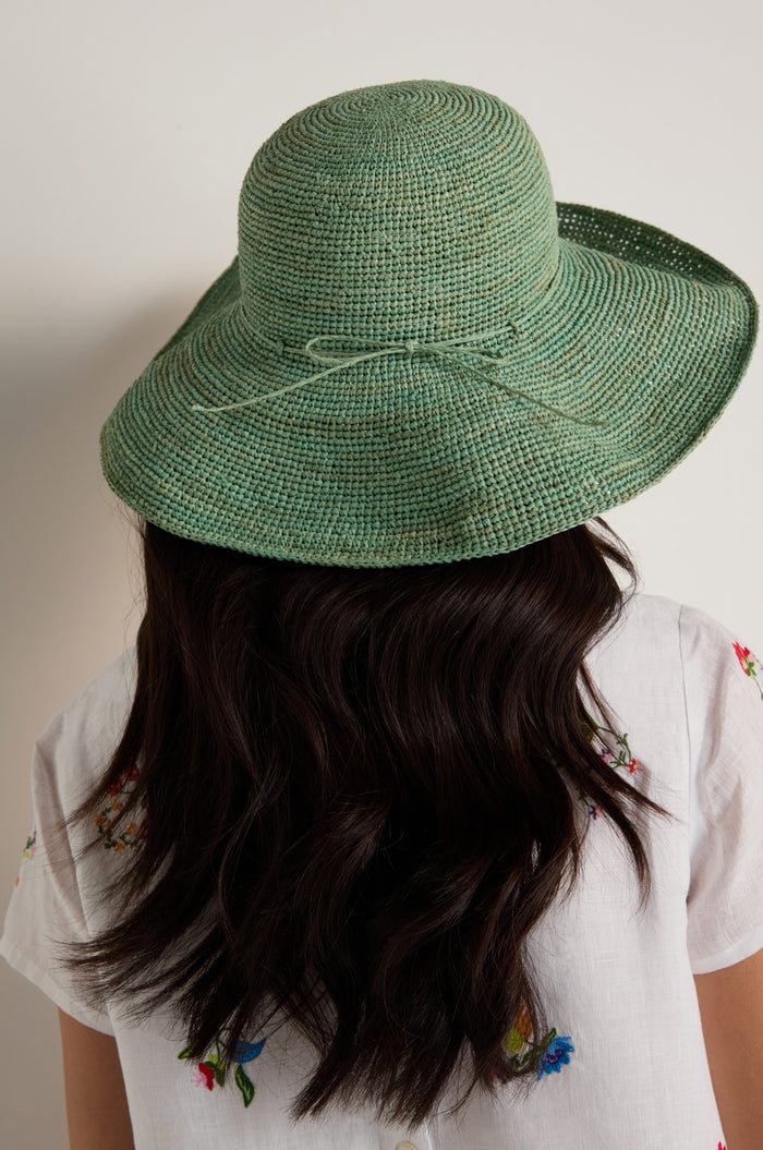 A person with long dark hair, seen from behind, wears a Handwoven Wide Brim Raffia Hat and a white shirt with colorful floral embroidery.