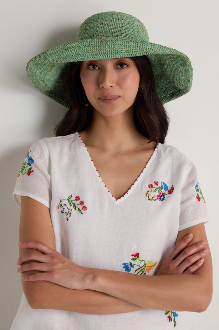 A woman wearing the Handwoven Wide Brim Raffia Hat and a white V-neck blouse with colorful floral embroidery stands with her arms crossed against a plain background.
