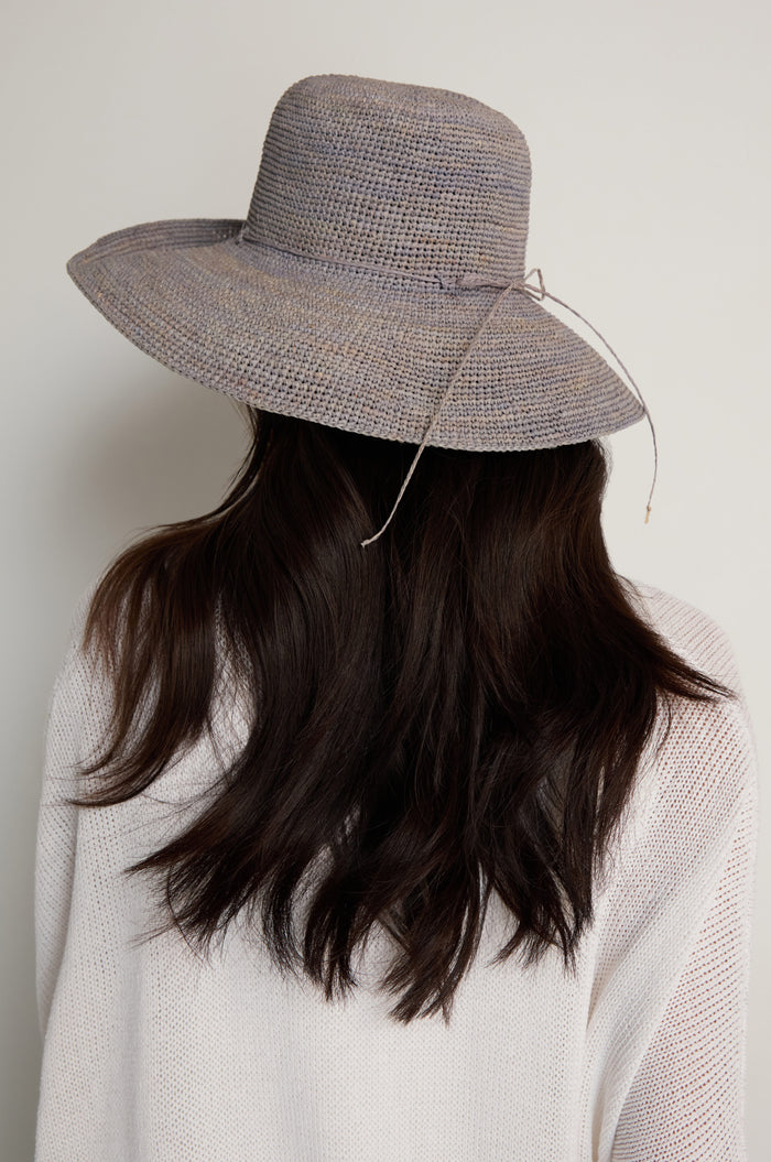 A person with long dark hair is shown from behind, wearing the Handwoven Wide Brim Raffia Hat and a light-colored sweater against a plain background.