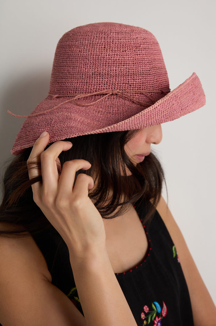A woman wearing the Handwoven Wide Brim Raffia Hat in pink and a black sleeveless floral top stands sideways, gently touching the brim of her hat.