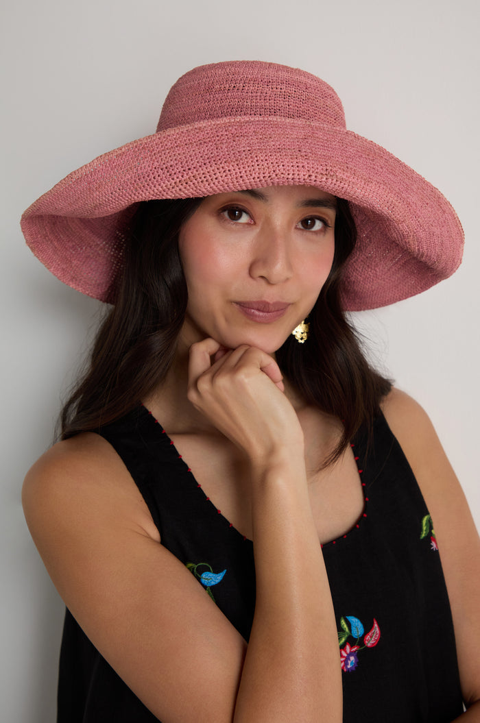 A woman poses against a plain background, wearing a black sleeveless top and the Handwoven Wide Brim Raffia Hat in pink, with one hand resting thoughtfully under her chin.