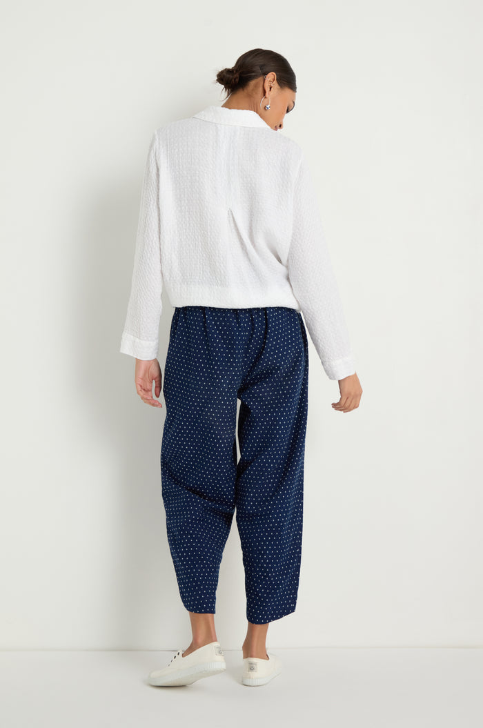 A woman stands against a white wall, facing away, wearing a white textured long-sleeve top and the Indigo Spot Cotton Bubble Trouser with an elasticated waist and cropped bubble silhouette, paired with white sneakers.
