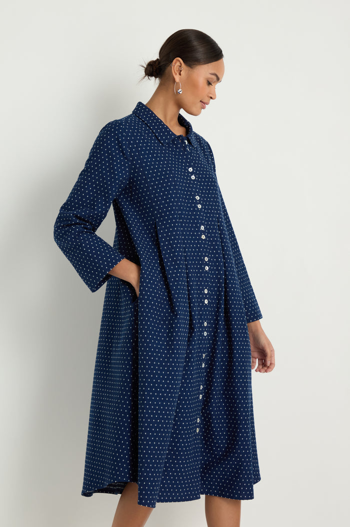 A woman models the Indigo Spot Cotton Shirt Dress, a long-sleeved, knee-length navy cotton dress with white polka dots and front buttons, highlighting its relaxed fit and subtle A-line silhouette against a white background.